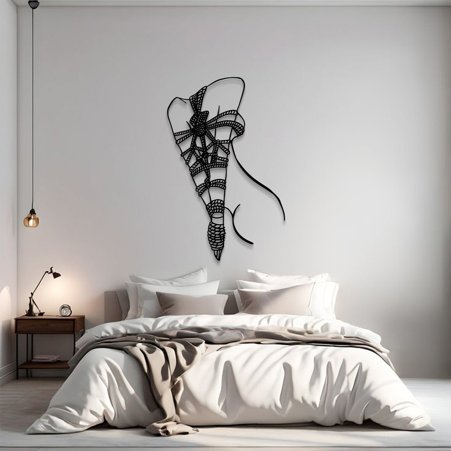 A modern bedroom scene with a bed dressed in white and beige bedding, a small wooden nightstand with a lamp, and a black wire wall art piece shaped like a stylized ribbon bow on the wall above the bed.