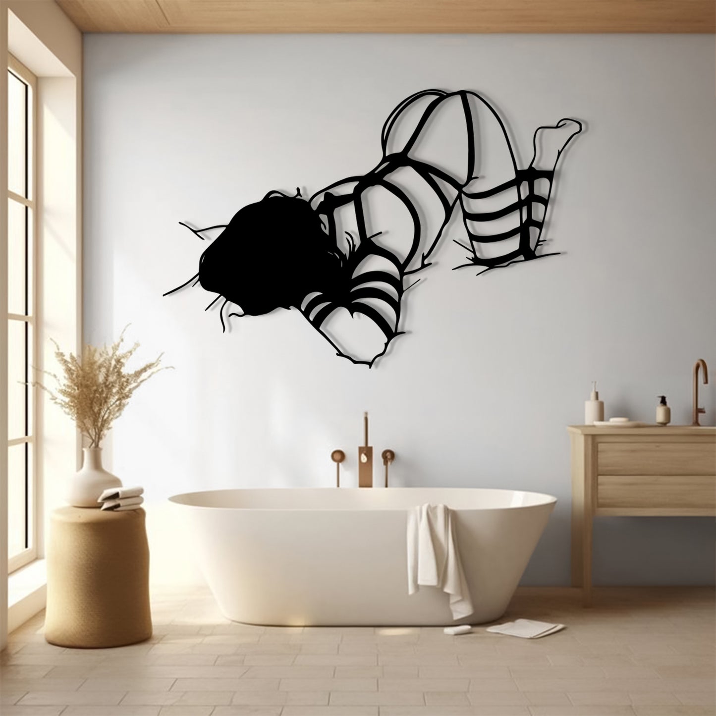A black metal wall art piece depicting a woman in strappy lingerie, displayed on a light gray wall above a white freestanding bathtub in a modern, sunlit bathroom.