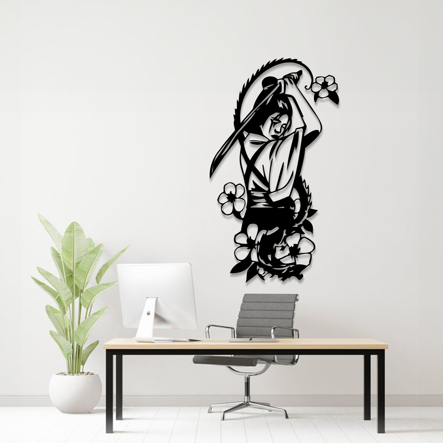 A black metal wall art piece depicting a woman with a sword and surrounding flowers, mounted on a white wall above a modern desk setup with a computer, chair, and potted plant.