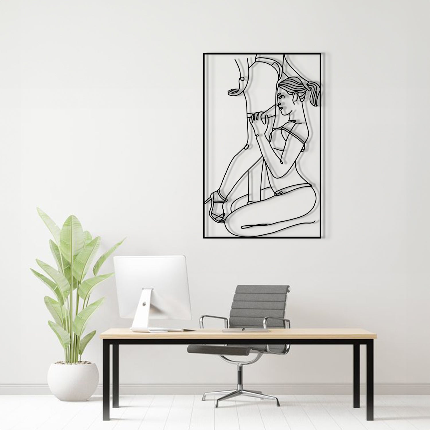 A black and white line art poster of a woman in a bikini hanging on a light gray wall above a modern desk setup with a computer, chair, and a potted plant.