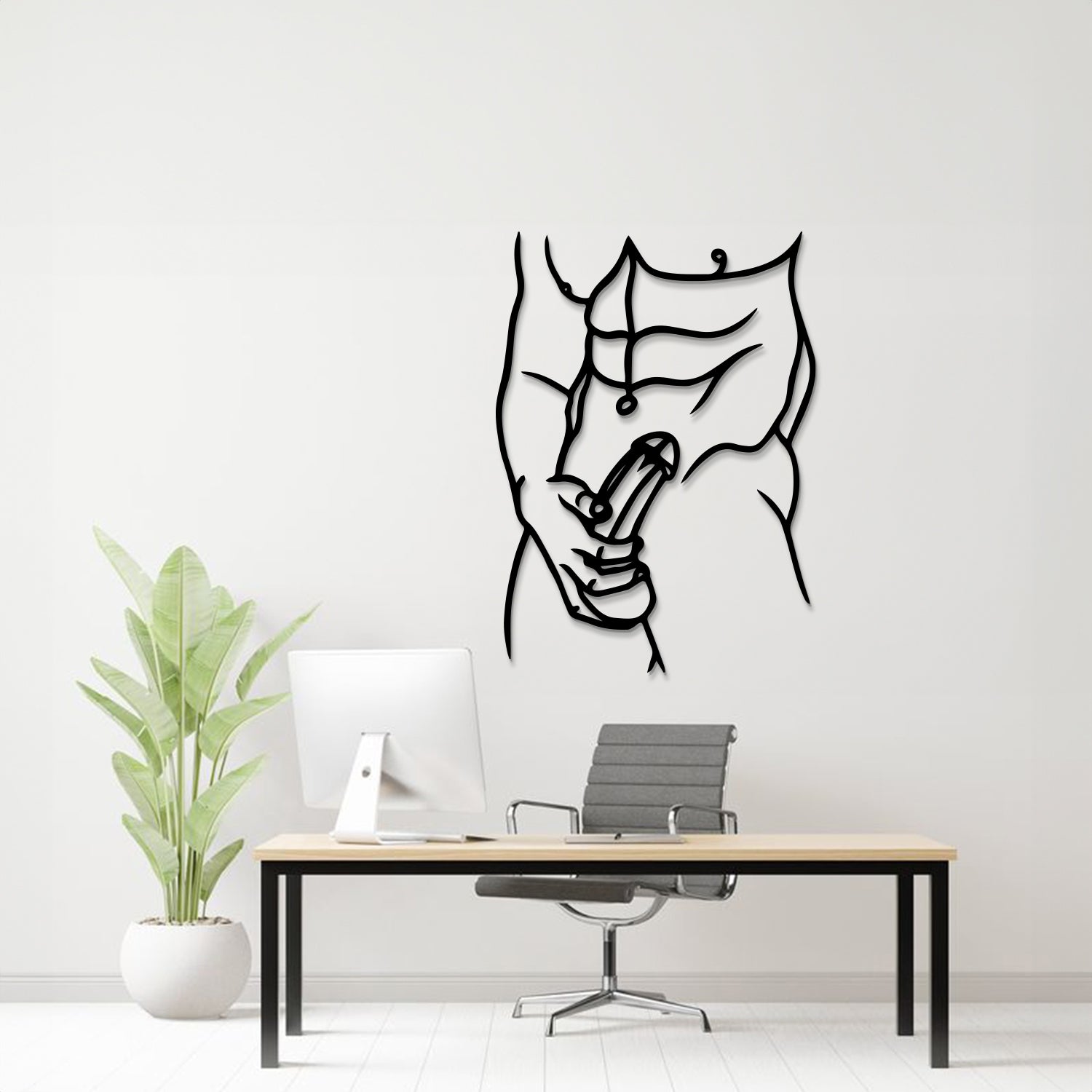 A minimalist black metal wall art piece depicting a muscular torso and hand holding a penis, displayed on a white wall above a modern desk setup with a computer, chair, and potted plant.