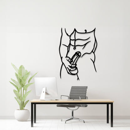 A minimalist black metal wall art piece depicting a muscular torso and hand holding a penis, displayed on a white wall above a modern desk setup with a computer, chair, and potted plant.