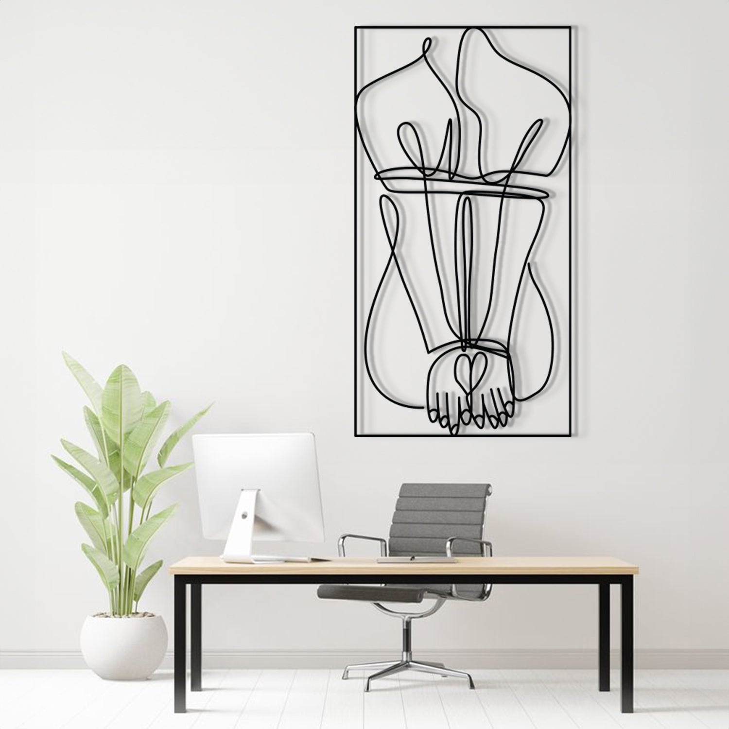 A minimalist black and white continuous line art print of hands holding hearts, displayed on a white wall above a modern desk setup with a plant and office chair.