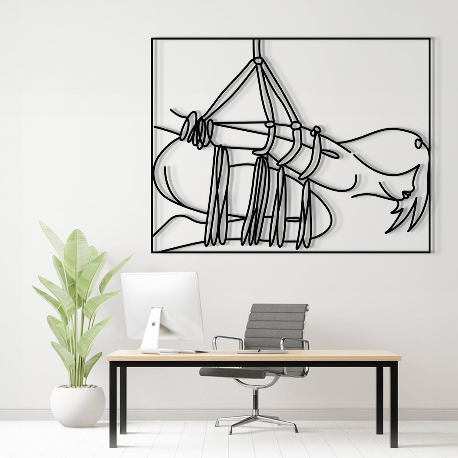 A minimalist black line art print of a hammock hanging over a reclining figure, displayed on a white wall above a modern desk setup with a computer, chair, and potted plant.