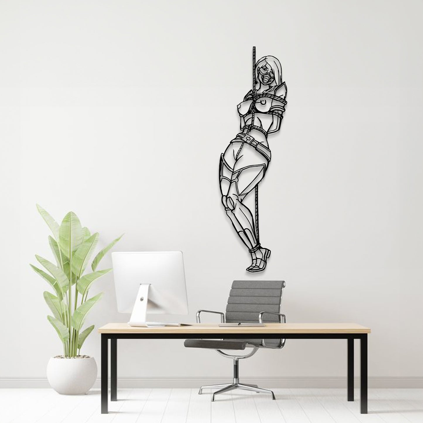 A black metal wall art piece depicting a bound woman hanging from a pole, displayed on a light gray wall above a modern desk setup with a computer, chair, and potted plant.