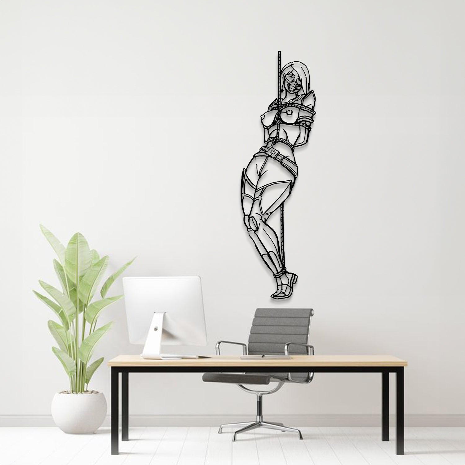 A black metal wall art piece depicting a bound woman hanging from a pole, displayed on a light gray wall above a modern desk setup with a computer, chair, and potted plant.