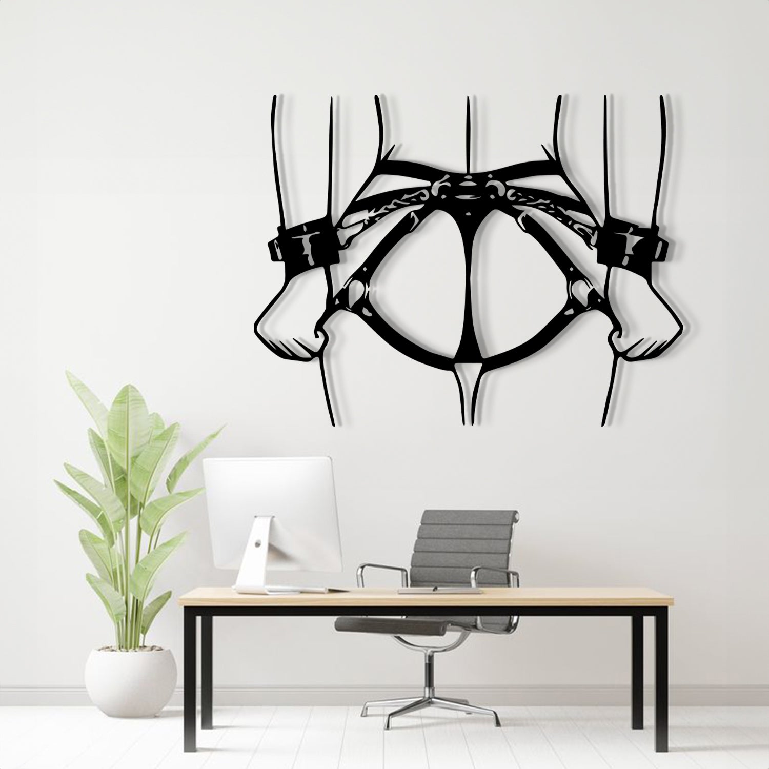 Black metal wall art depicting a harness and hands, displayed on a white wall above a modern desk setup with a computer, chair, and potted plant.