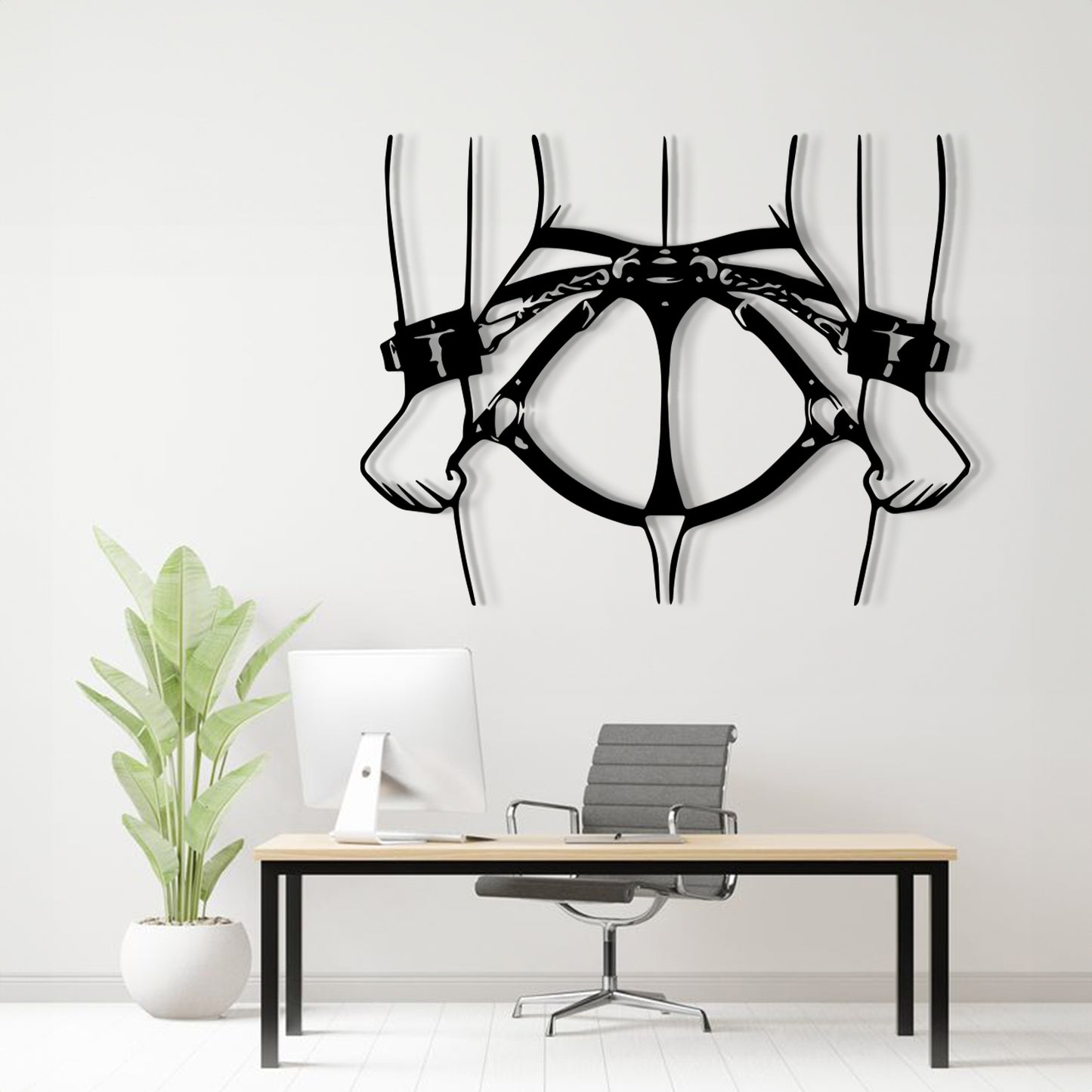 Black metal wall art depicting a harness and hands, displayed on a white wall above a modern desk setup with a computer, chair, and potted plant.
