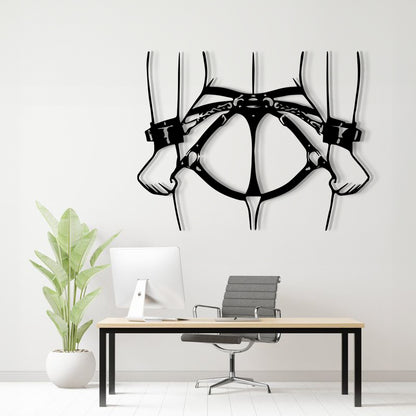 Black metal wall art depicting a harness and hands, displayed on a white wall above a modern desk setup with a computer, chair, and potted plant.