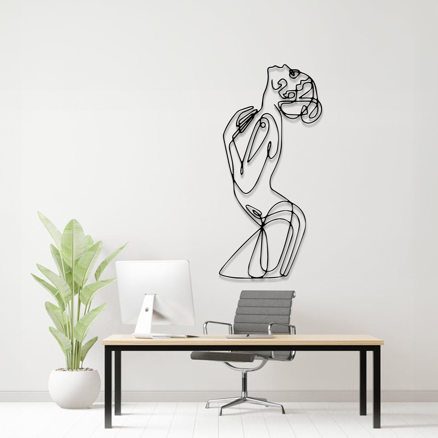A minimalist black metal line art wall sculpture of a woman in a flowing dress, mounted on a white wall above a modern desk setup with a computer, chair, and potted plant.