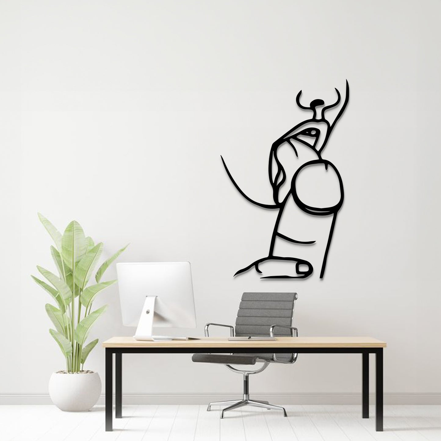 A minimalist black line art wall decal of a seated figure with horns, displayed on a light gray wall above a modern desk setup with a computer, chair, and potted plant.