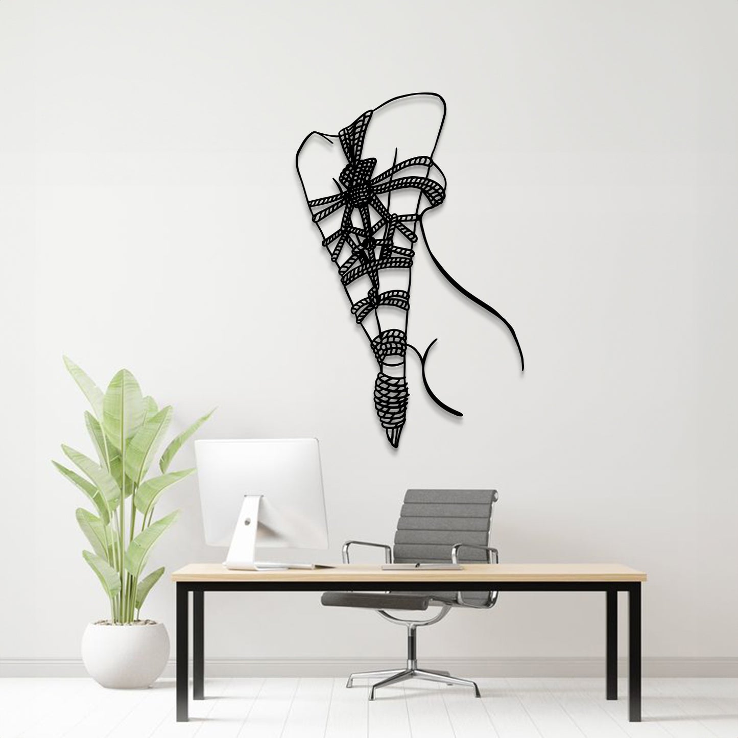 A black metal wall art sculpture of a stylized bow and arrow hangs on a light gray wall above a modern desk setup with a computer monitor, chair, and potted plant.