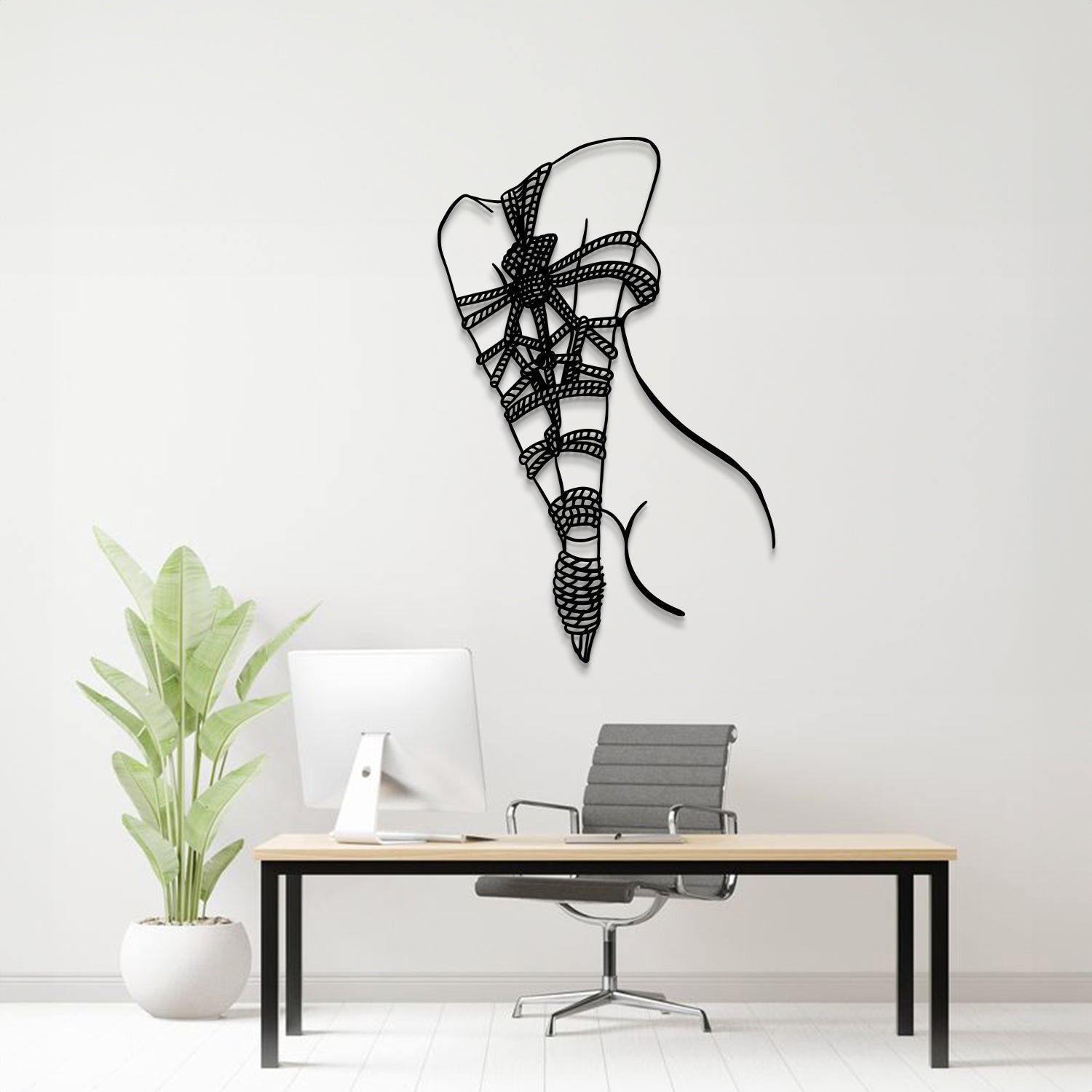 A black metal wall art sculpture of a stylized bow and arrow hangs on a light gray wall above a modern desk setup with a computer monitor, chair, and potted plant.