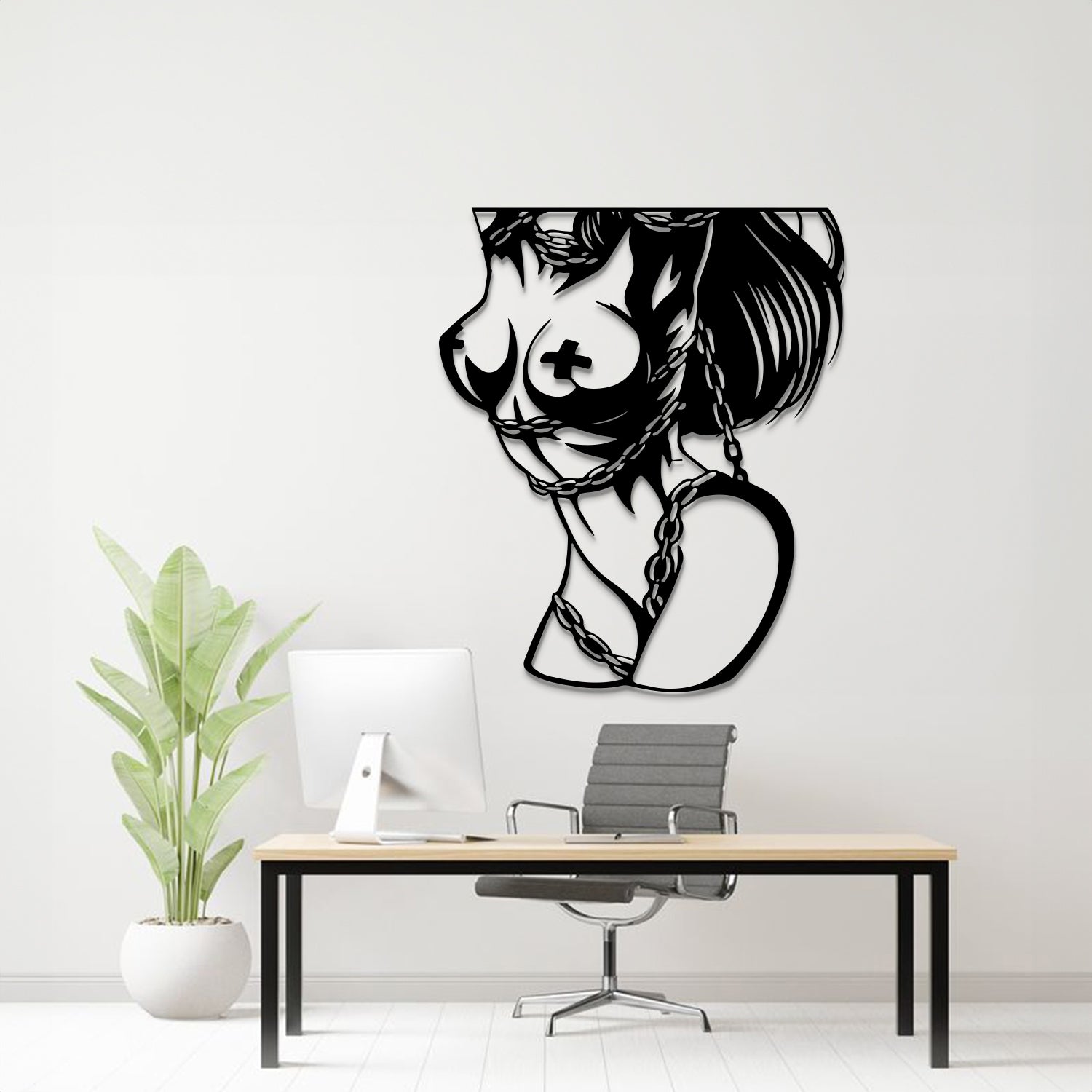 A black metal wall art piece depicting a stylized female profile with a cross on her breast and a heart-shaped chain motif, displayed on a white wall above a modern desk setup with a computer, chair, and potted plant.