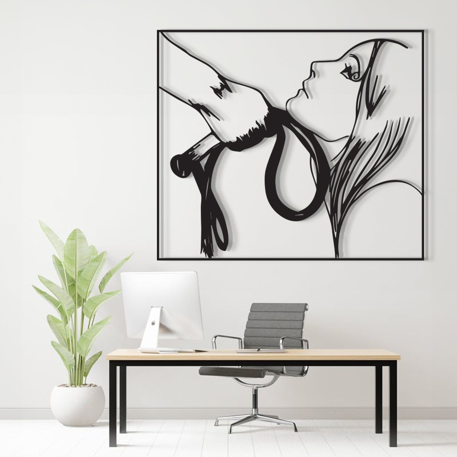 A large black and white framed wall art print depicting a stylized hand holding a rope, hanging above a modern desk setup with a computer, chair, and potted plant in a bright office setting.