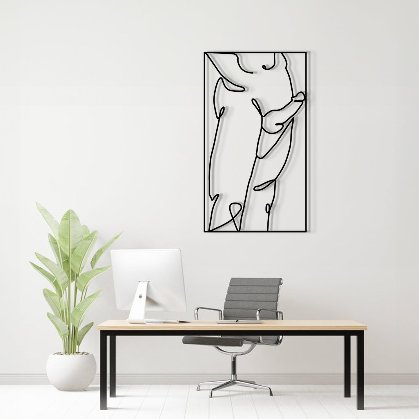 A minimalist black line art metal wall art print hangs on a white wall above a modern desk setup with a computer, office chair, and a potted plant.