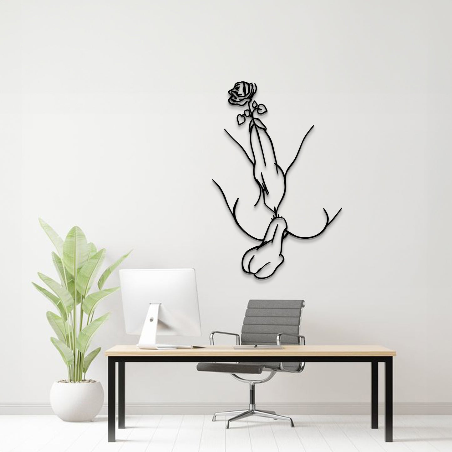 A minimalist black metal wall art piece depicting a hand holding a rose, mounted on a white wall above a modern desk setup with a computer, chair, and potted plant.