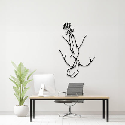 A minimalist black metal wall art piece depicting a hand holding a rose, mounted on a white wall above a modern desk setup with a computer, chair, and potted plant.