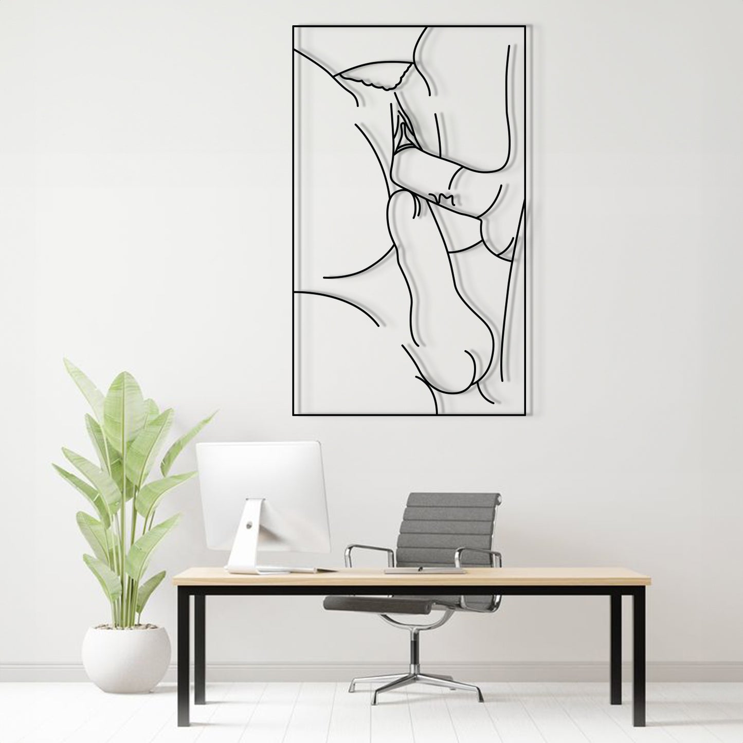 A minimalist black and white continuous line art print of a crouching figure, displayed on a white wall above a modern desk setup with a computer, chair, and potted plant.
