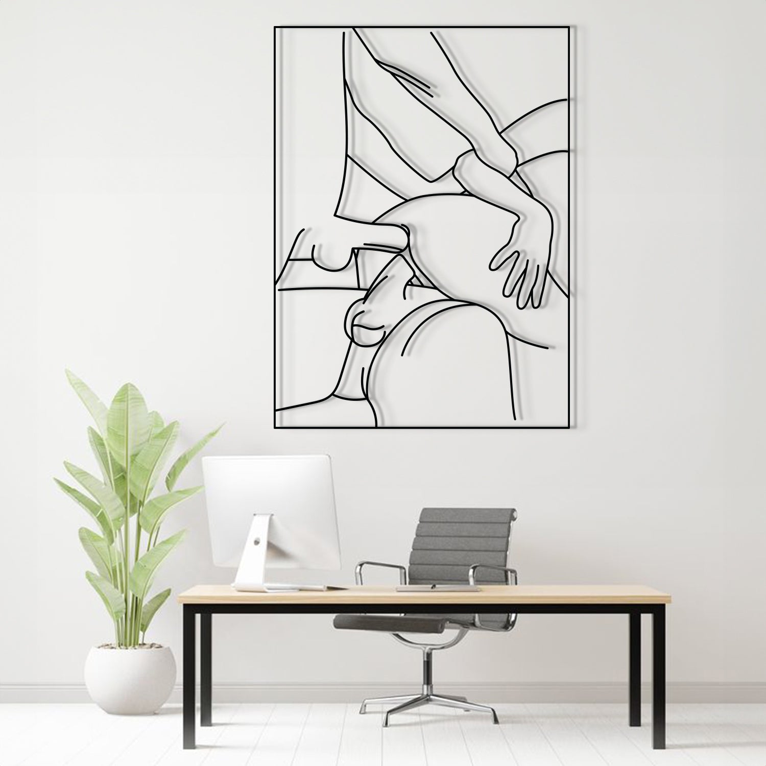 A minimalist black and white continuous line art print of a massage or touch-up scene, displayed on a light gray wall above a modern desk setup with a computer, office chair, and a potted plant.