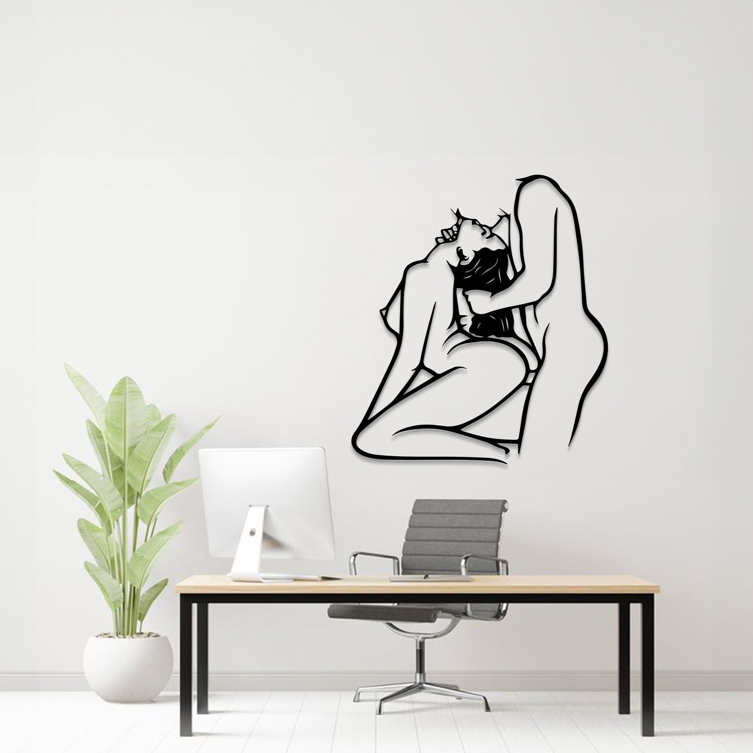 A minimalist black line art wall decal of an embracing couple is displayed on a white wall in a modern home office setting, above a desk with a computer, chair, and a potted plant.