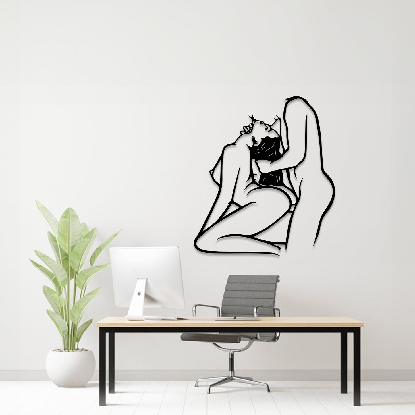 A minimalist black line art wall decal of an embracing couple is displayed on a white wall in a modern home office setting, above a desk with a computer, chair, and a potted plant.