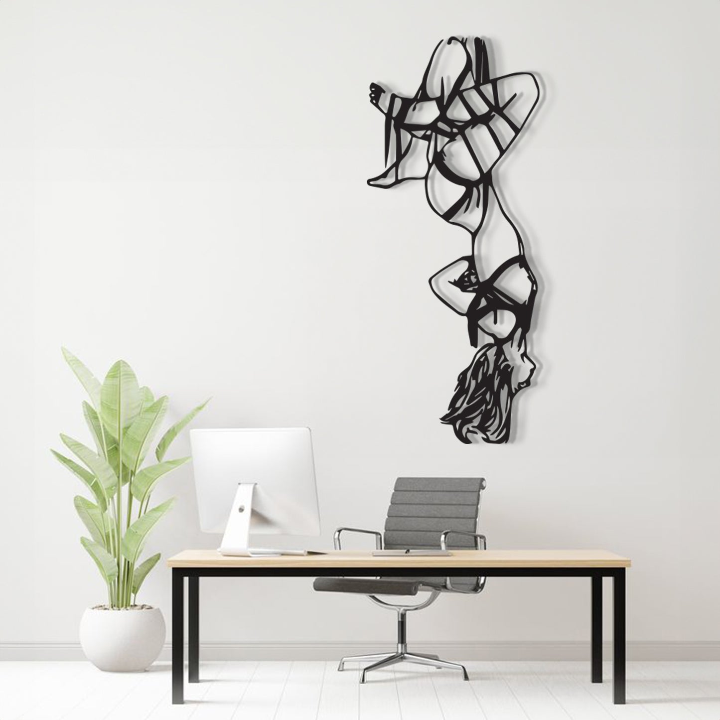 A black metal wall art sculpture of a stylized dancer hangs on a white wall above a minimalist desk setup with a computer, chair, and potted plant.