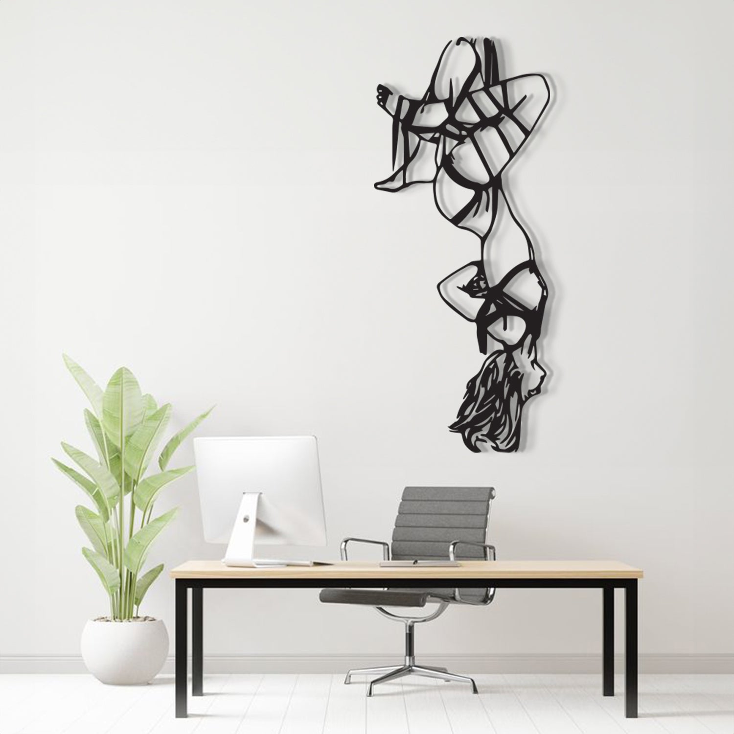 A black metal wall art sculpture of a stylized dancer hangs on a white wall above a minimalist desk setup with a computer, chair, and potted plant.