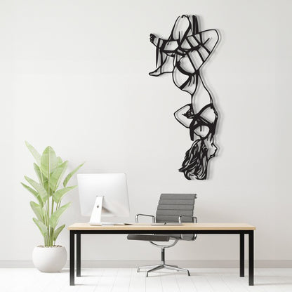 A black metal wall art sculpture of a stylized dancer hangs on a white wall above a minimalist desk setup with a computer, chair, and potted plant.