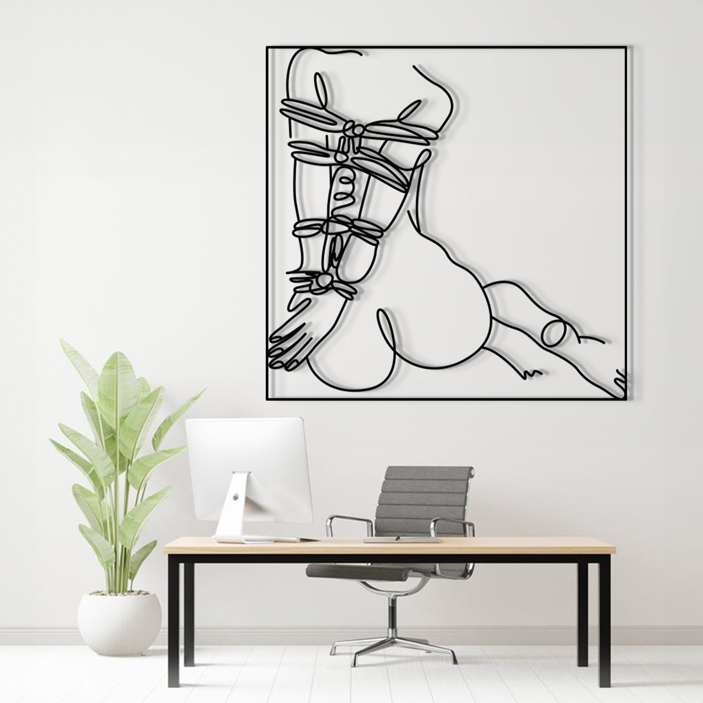 A minimalist black and white continuous line art print of a woman's body, displayed on a white wall above a modern desk setup with a computer, chair, and potted plant.