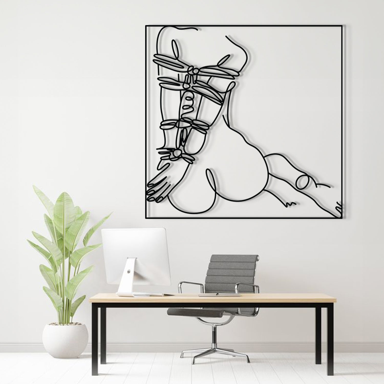 A minimalist black and white continuous line art print of a woman's body, displayed on a white wall above a modern desk setup with a computer, chair, and potted plant.