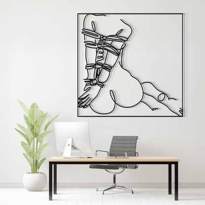A minimalist black and white continuous line art print of a woman's body, displayed on a white wall above a modern desk setup with a computer, chair, and potted plant.