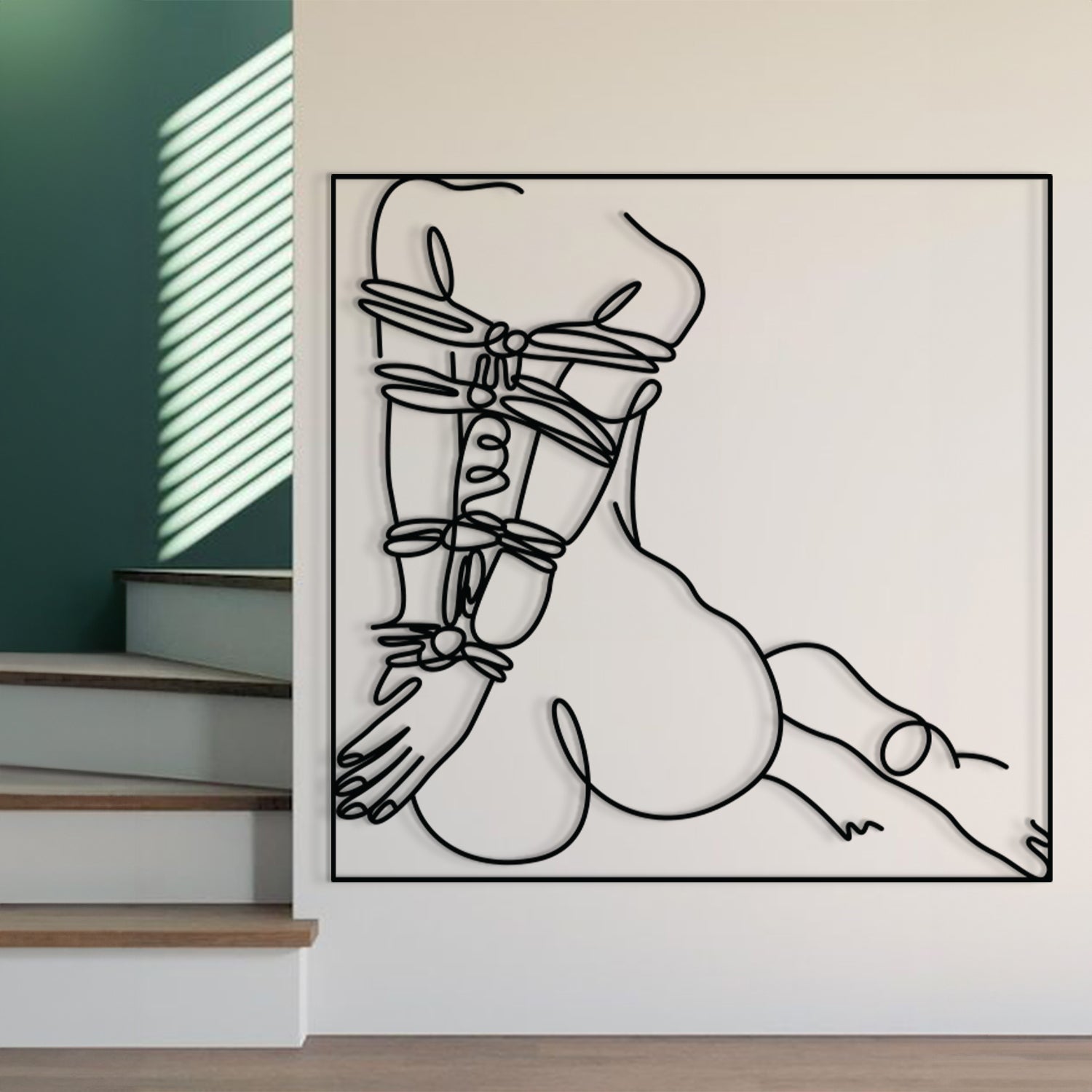 A minimalist black and white continuous line art print of a kneeling nude female figure, displayed on a white wall in a modern interior setting with a staircase and green accent wall visible.
