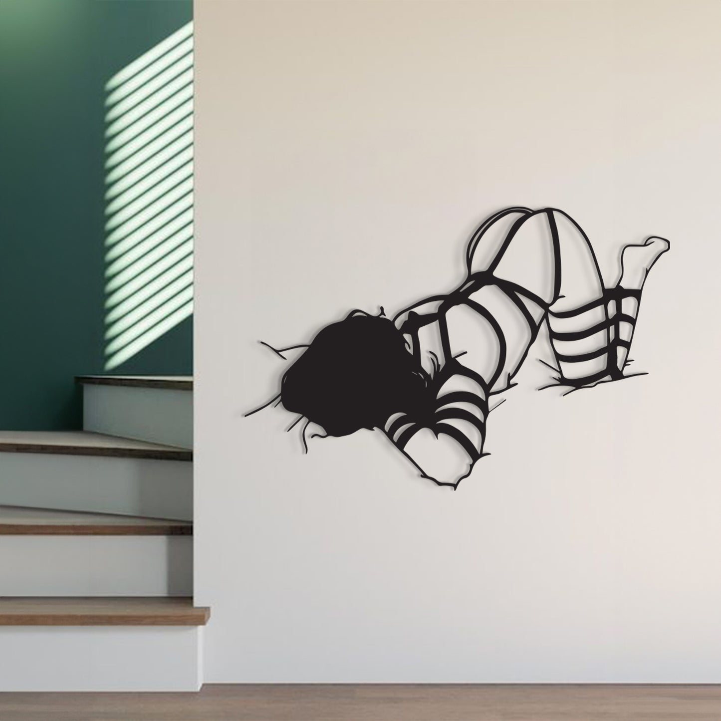 A black metal wall art piece depicting a woman in a strappy dress breaking through a wall, shown mounted on a white interior wall next to a staircase.