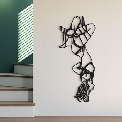 A black metal wall art sculpture depicting a stylized female figure in a strappy dress, shown mounted on a white wall in a modern interior setting with stairs and a green accent wall visible.