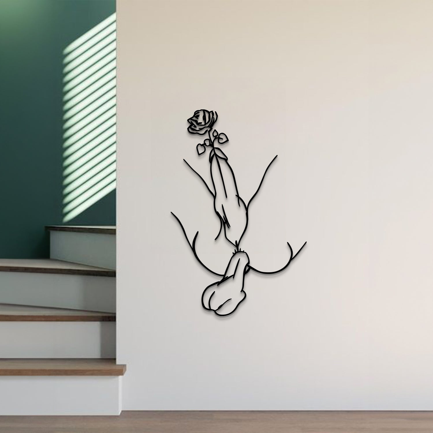 A minimalist black metal wall art piece depicting a hand holding a rose, mounted on a light beige wall in a modern interior setting with wooden stairs and green walls visible.