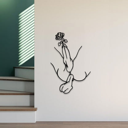 A minimalist black metal wall art piece depicting a hand holding a rose, mounted on a light beige wall in a modern interior setting with wooden stairs and green walls visible.