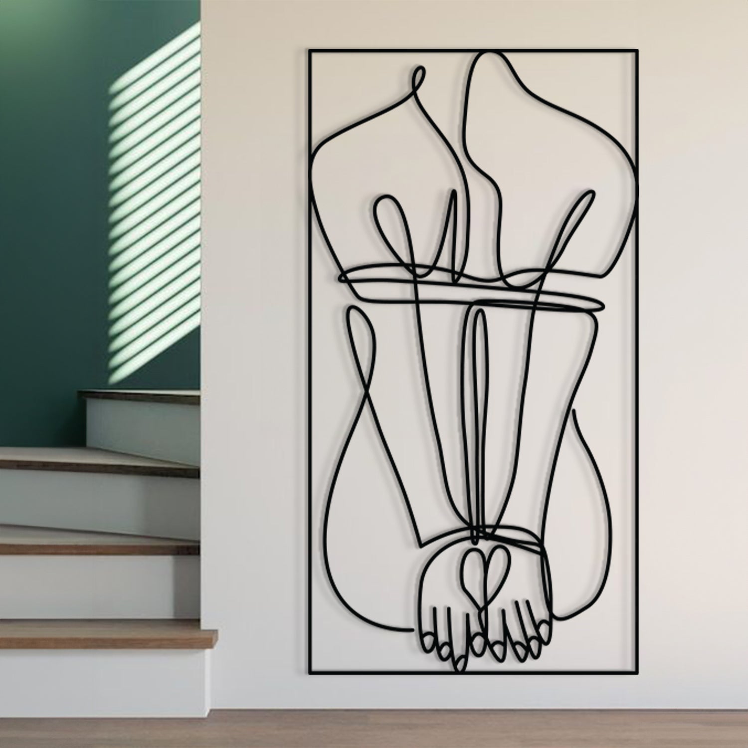 A minimalist black and white continuous line wall art print depicting a heart-shaped hand and legs, displayed on a white wall in a modern interior setting next to a staircase.