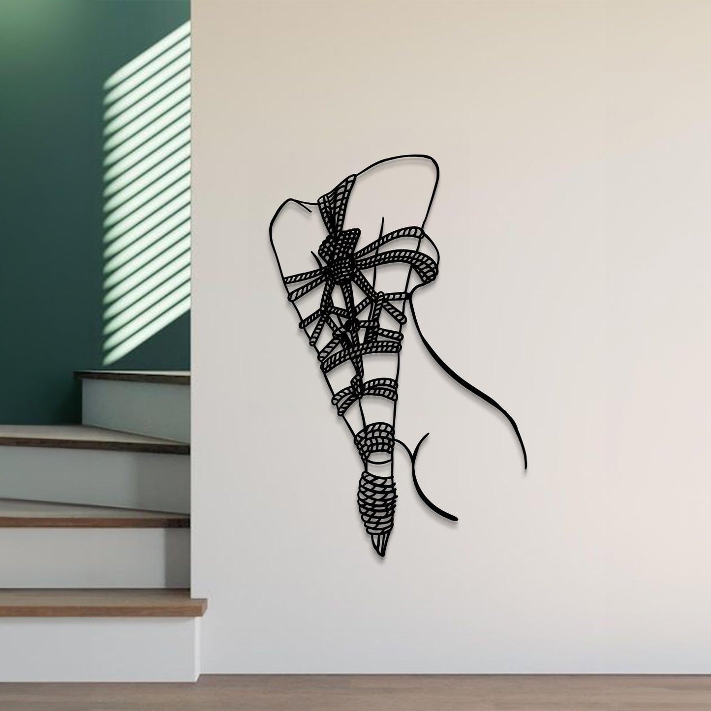 A black metal wall art sculpture of a lace-up high-heel shoe is mounted on a white wall in a modern interior setting with a staircase and green accent wall visible.