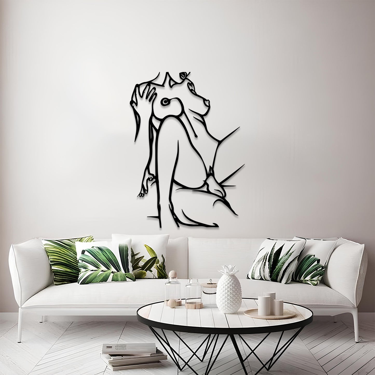 A modern living room scene featuring a black metal wall art piece of a seated figure in profile mounted on a light gray wall above a white sofa with green leaf-patterned pillows.