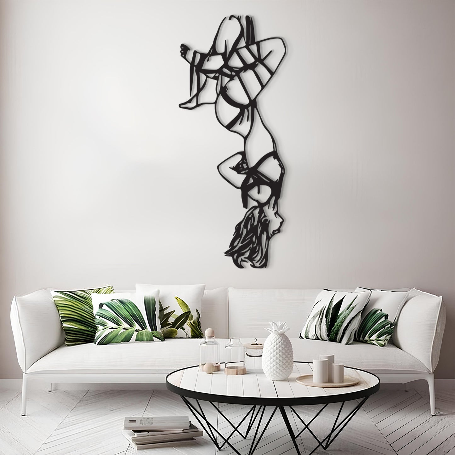A modern living room scene featuring a white sofa with green tropical leaf patterned throw pillows, a round black coffee table, and a tall black metal abstract wall sculpture mounted on a light gray wall.