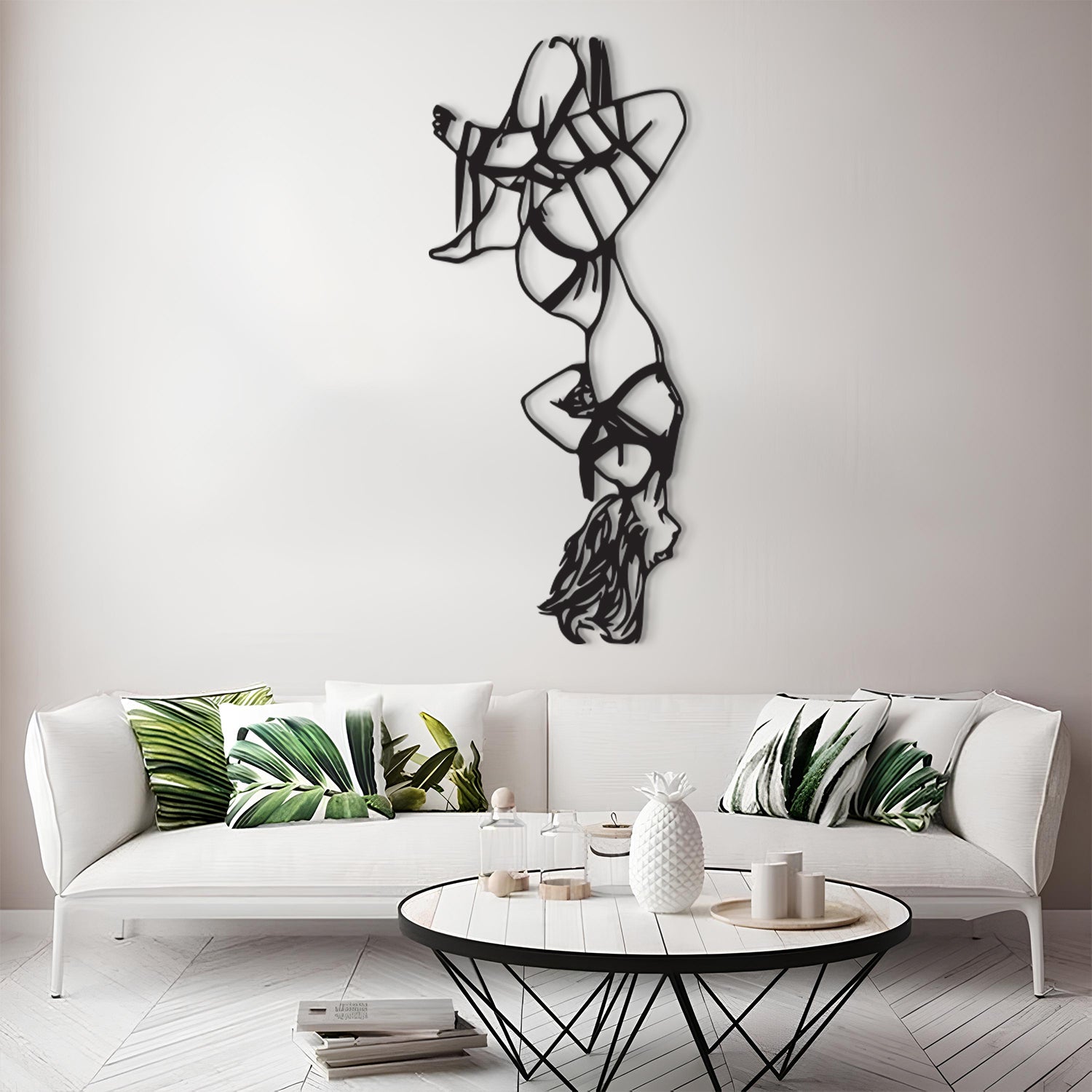 A modern living room scene featuring a white sofa with green tropical leaf patterned throw pillows, a round black coffee table, and a tall black metal abstract wall sculpture mounted on a light gray wall.
