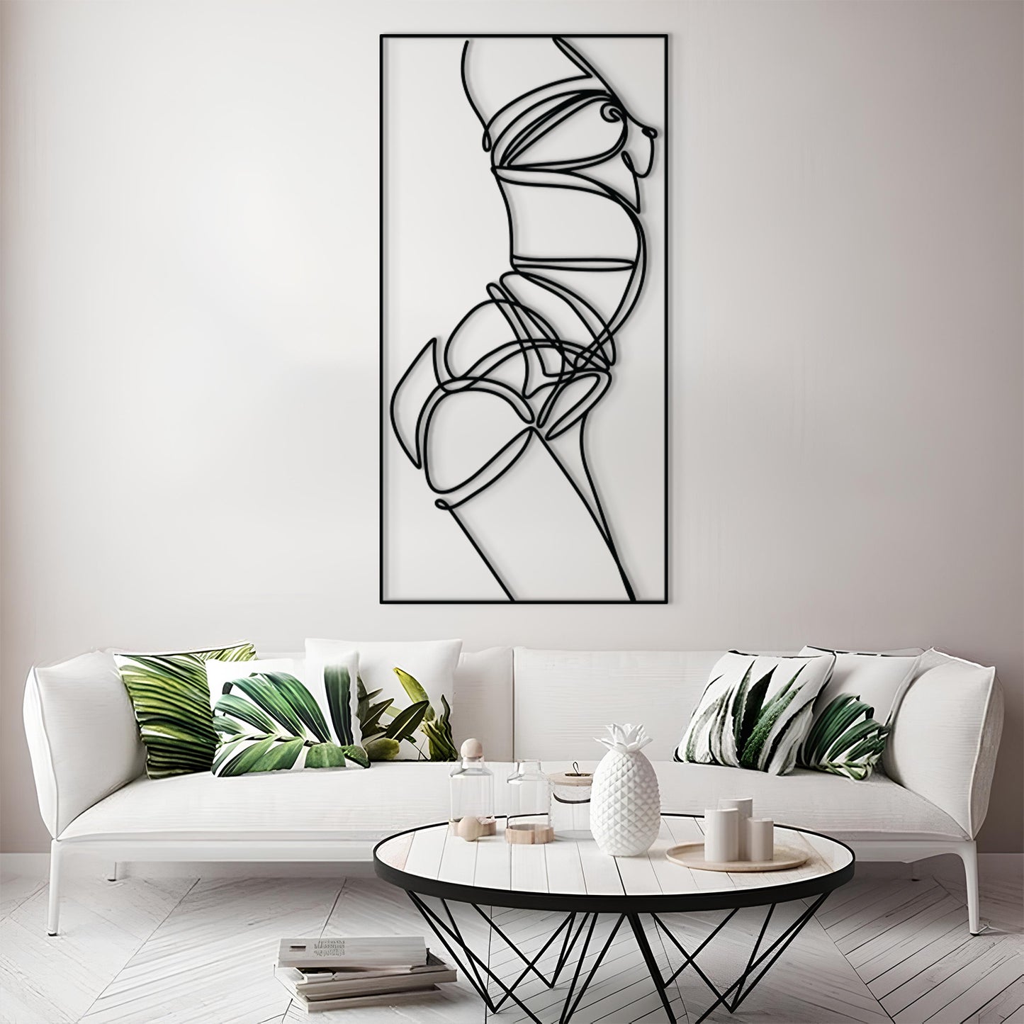 A modern living room scene featuring a tall black-and-white minimalist line art wall print above a white sofa with green leaf-patterned pillows. A round coffee table sits in front of the sofa on a light wood floor.