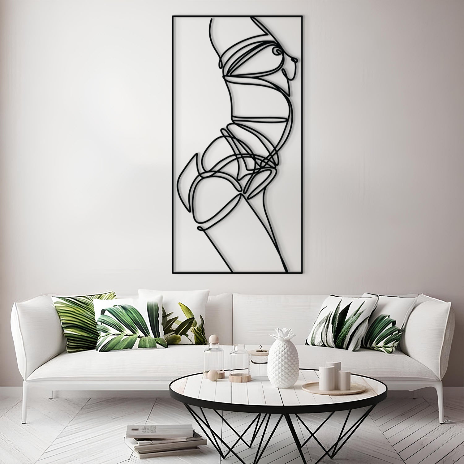 A modern living room scene featuring a tall black-and-white minimalist line art wall print above a white sofa with green leaf-patterned pillows. A round coffee table sits in front of the sofa on a light wood floor.