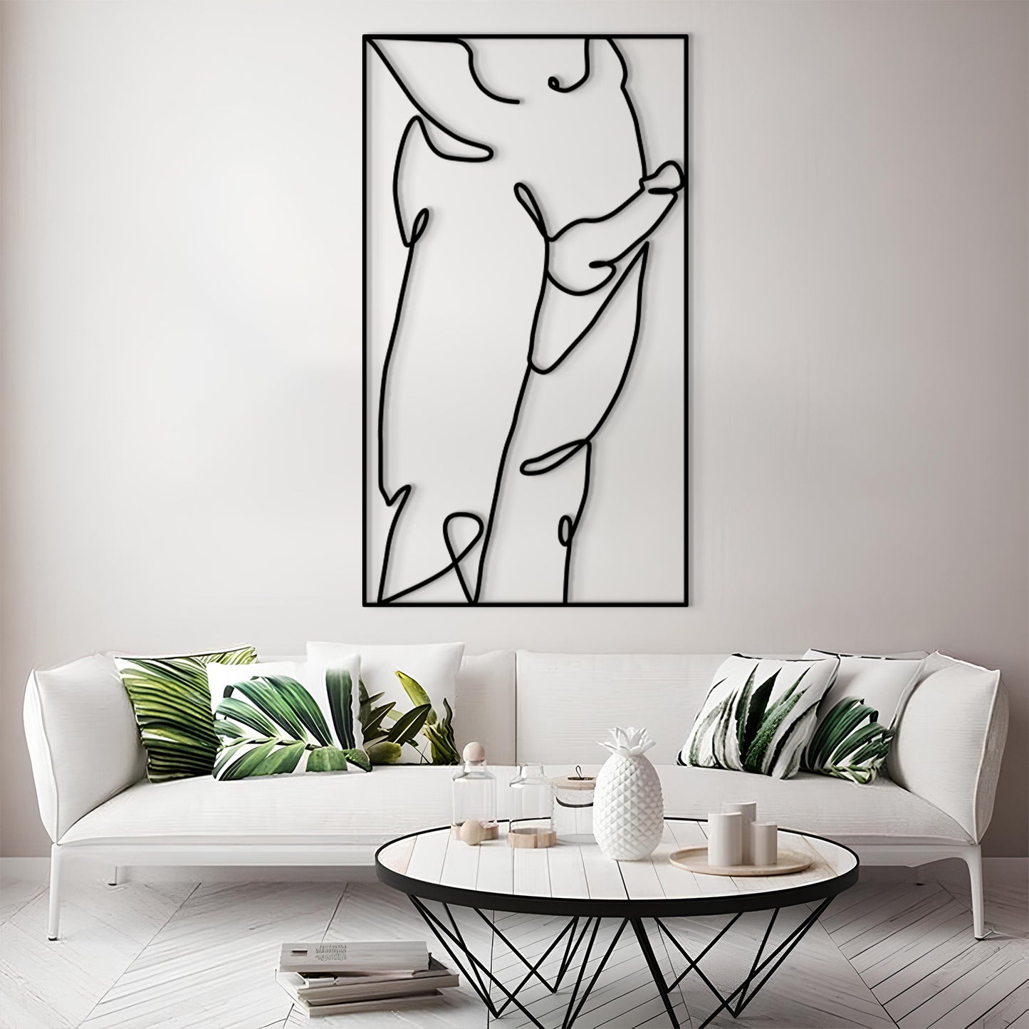 A modern living room scene featuring a large black and white minimalist line art print on a white wall above a white sofa with green leaf-patterned pillows. In the foreground is a round white coffee table with a black wire frame.