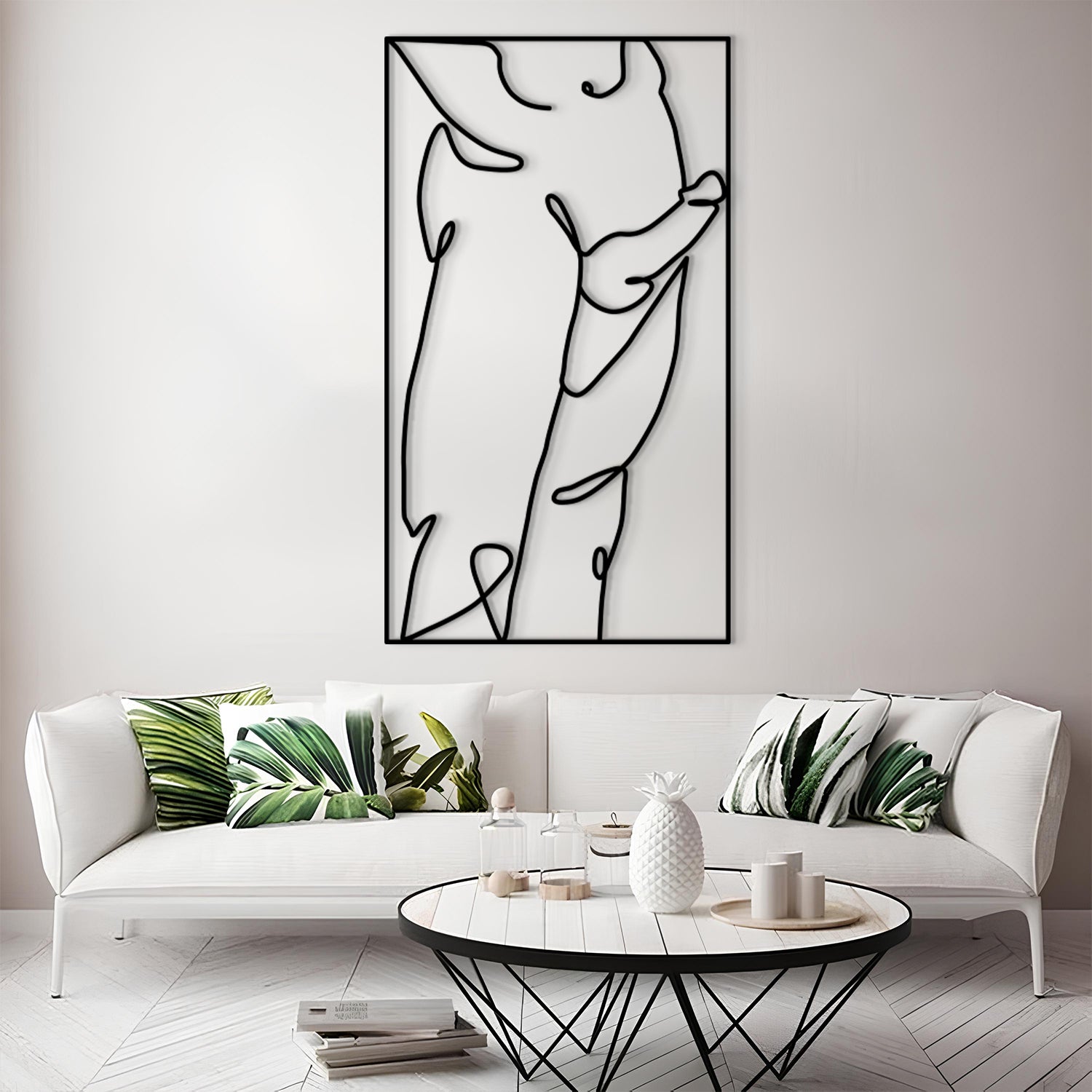 A modern living room scene featuring a large black and white minimalist line art print on a white wall above a white sofa with green leaf-patterned pillows. In the foreground is a round white coffee table with a black wire frame.