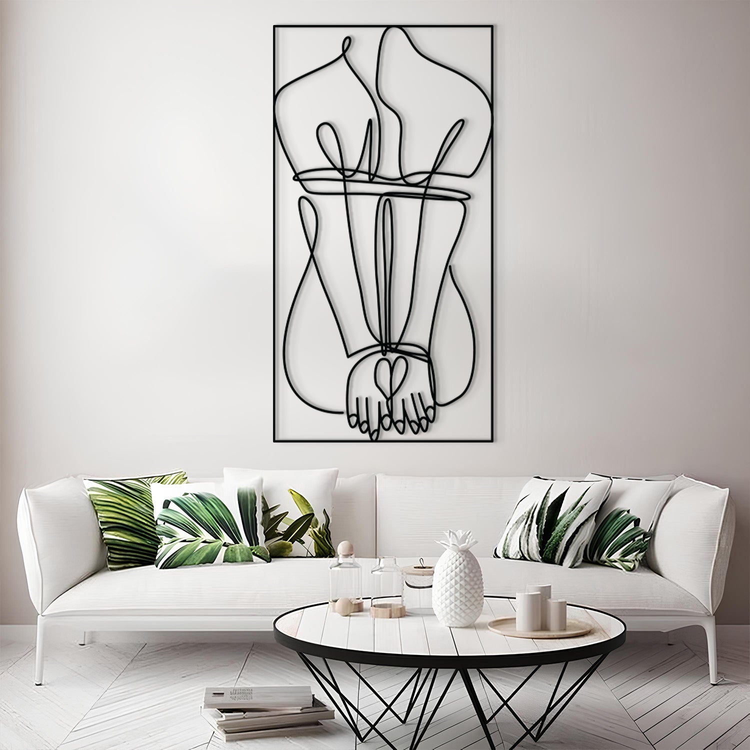 A modern living room scene featuring a large black wire art piece of intertwined hands hanging on a light gray wall above a white sofa with green leaf-patterned pillows.
