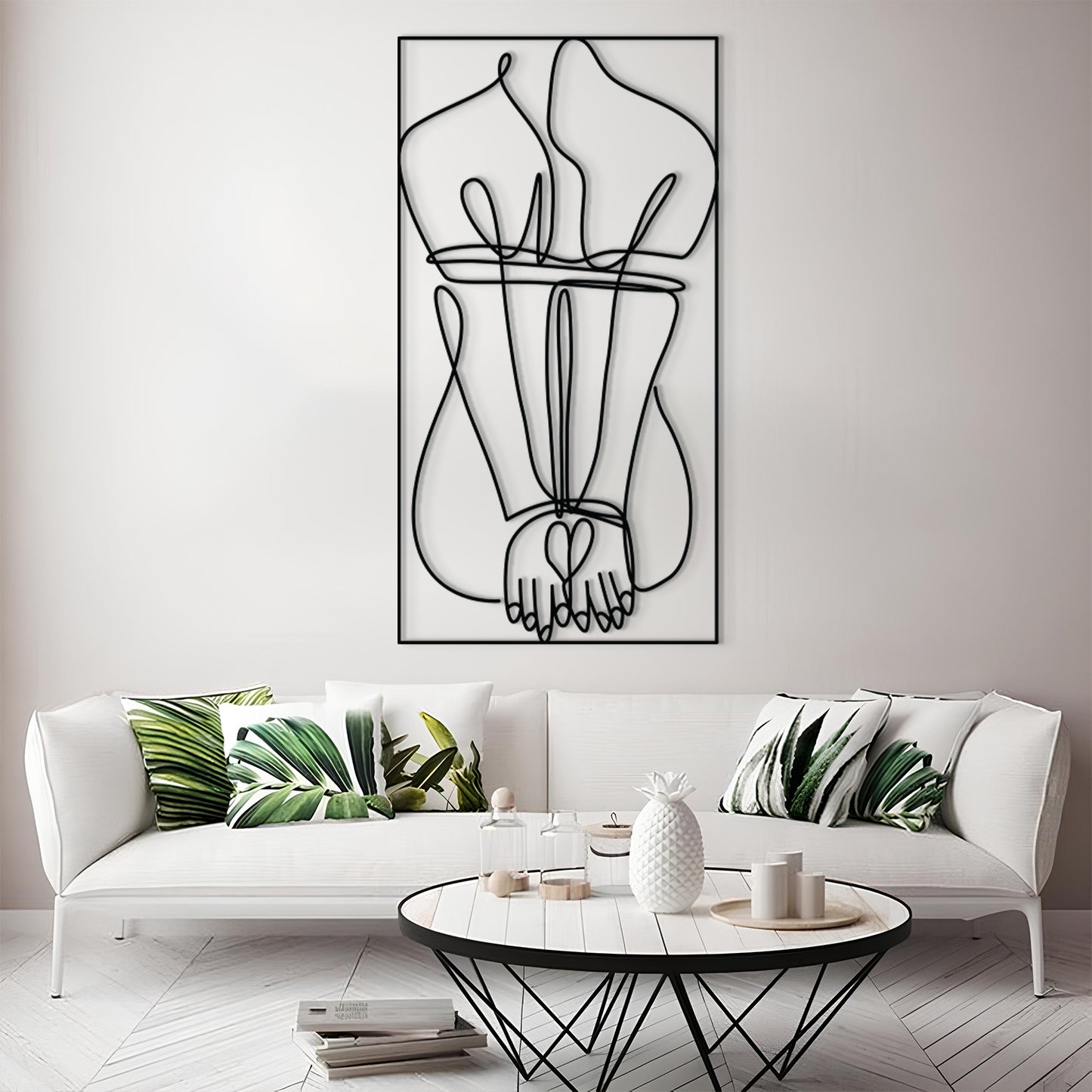 A modern living room scene featuring a large black wire art piece of intertwined hands hanging on a light gray wall above a white sofa with green leaf-patterned pillows.