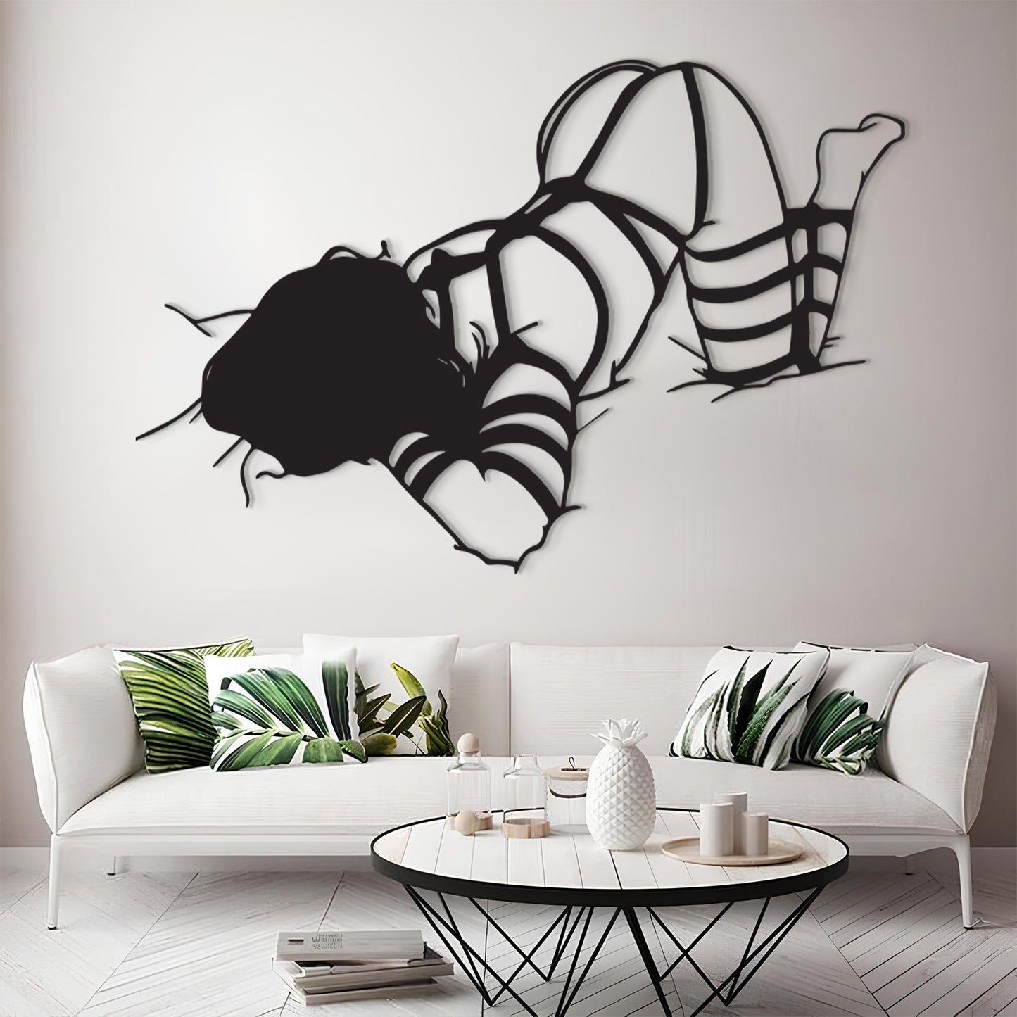 A black metal wall art piece depicting a woman in strappy high-heel sandals, displayed on a light gray wall above a modern white sofa with green tropical leaf pillows.