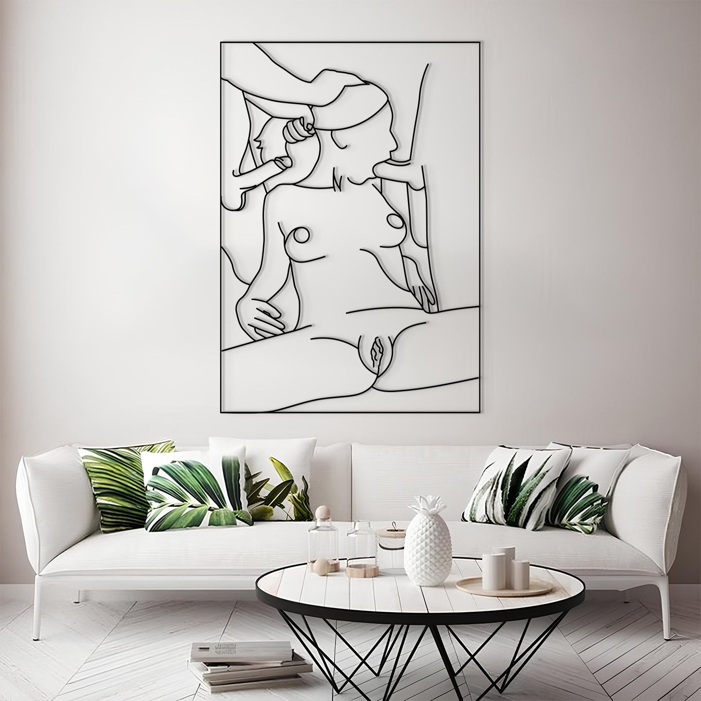 A framed minimalist black line art print of a nude female figure is displayed on a light gray wall above a white sofa with green leaf-patterned pillows in a modern living room setting.